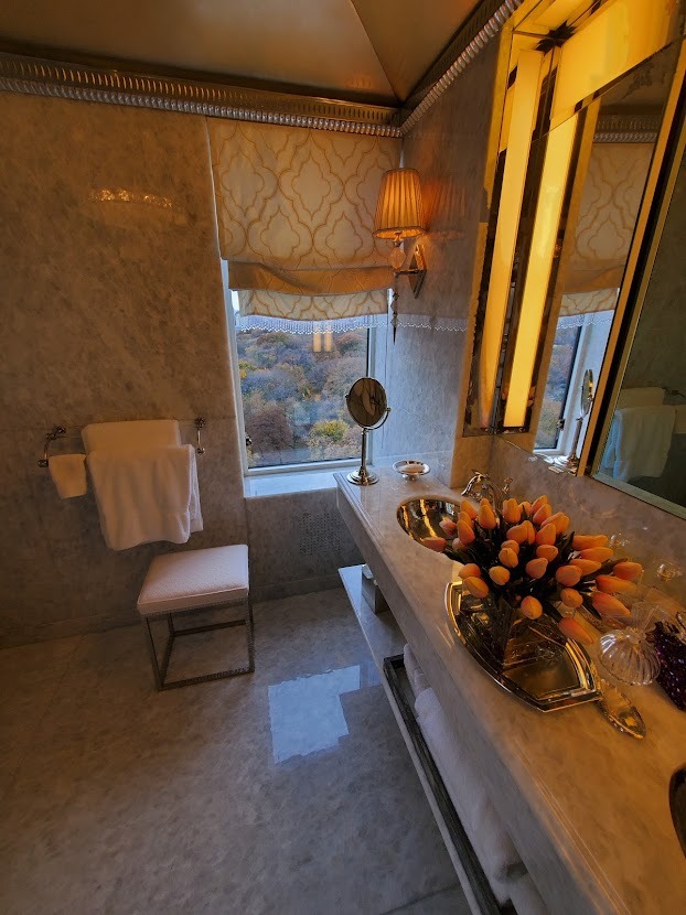 Elegancka łazienka z marmurowymi ścianami, dekoracyjnym lustrem z oświetleniem, widokiem na zalesiony krajobraz za oknem i bukietem pomarańczowych tulipanów na blacie umywalki.