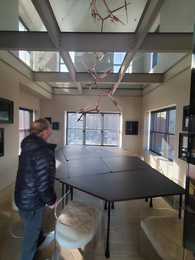 Duży, czarny stół konferencyjny z modułowym blatem w jasnym pomieszczeniu z oknami i przeszklonym dachem, oświetlonym przez nowoczesną lampę wiszącą.