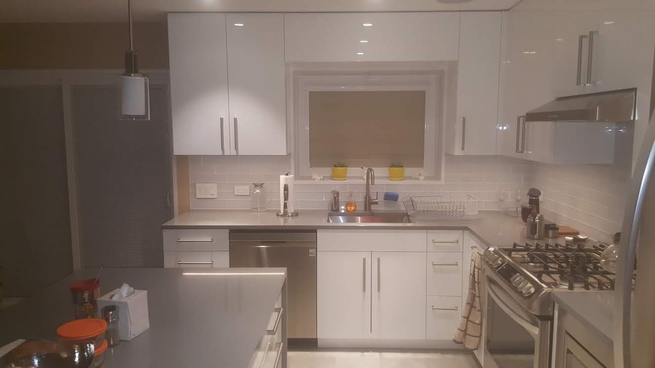 Biała kuchnia z połyskiem, szare blaty, płytki na ścianie, okno z doniczkami, nowoczesne oświetlenie wiszące, elementy wyposażenia AGD.