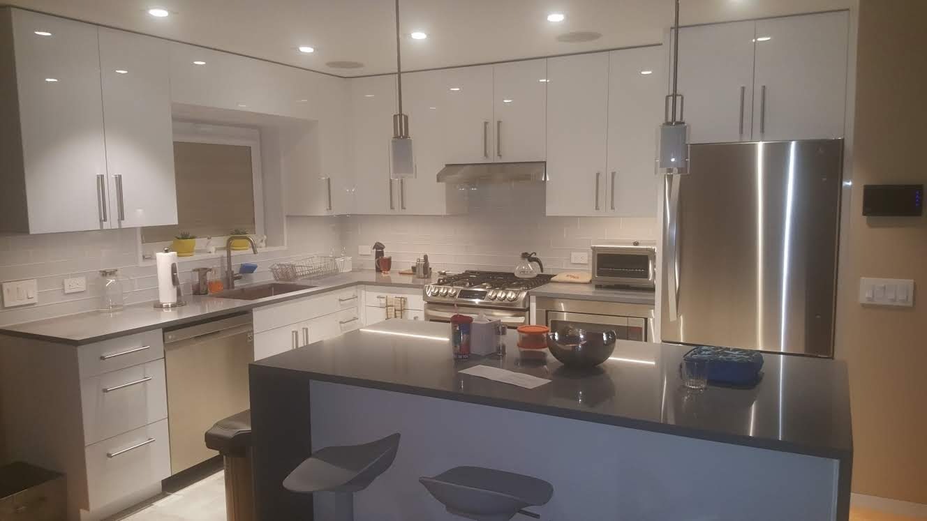 Biała kuchnia z połyskiem, wyspa kuchenna z szarym blatem, nowoczesne oświetlenie punktowe i wiszące lampy, stalowa lodówka i sprzęt AGD, widok na okno z roślinami.