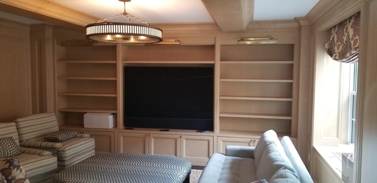 Jasna zabudowa ścienna z półkami i szafkami, centralnie umieszczony czarny telewizor, oświetlenie punktowe nad półkami, okrągła lampa sufitowa, fragment sofy i fotela w salonie.