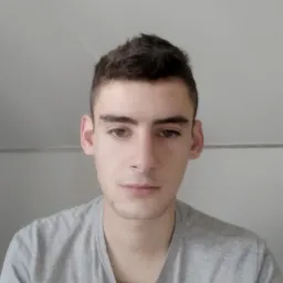 Portret młodego elektryka z Milicza w szarej koszulce, patrzącego prosto w obiektyw, na tle jasnej ściany.