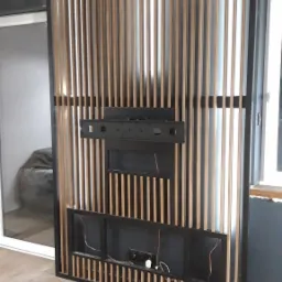 Dekoracyjny panel ażurowy z drewna i metalu, zamontowany na ścianie, z widocznymi otworami na kable i uchwytem na telewizor. Widoczne gniazdka elektryczne i wystające przewody.