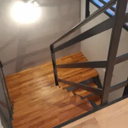 Metalowa poręcz w kolorze grafitowym, zamontowana przy drewnianych schodach z widocznym usłojeniem, oświetlona światłem z góry, w tle ściana w odcieniach szarości.