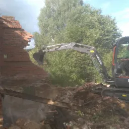 Mini koparka Volvo ECR25D usuwa fragment ceglanej ściany, widoczny operator w kabinie, w tle zieleń drzew.