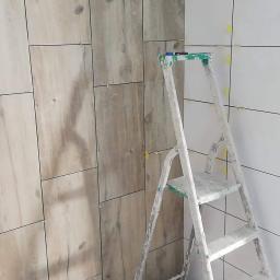 FH Enterprenor - Fragment łazienki w trakcie układania płytek imitujących drewno na ścianie, widoczna drabina malarska, poziomica i wiertarka udarowa Hilti.