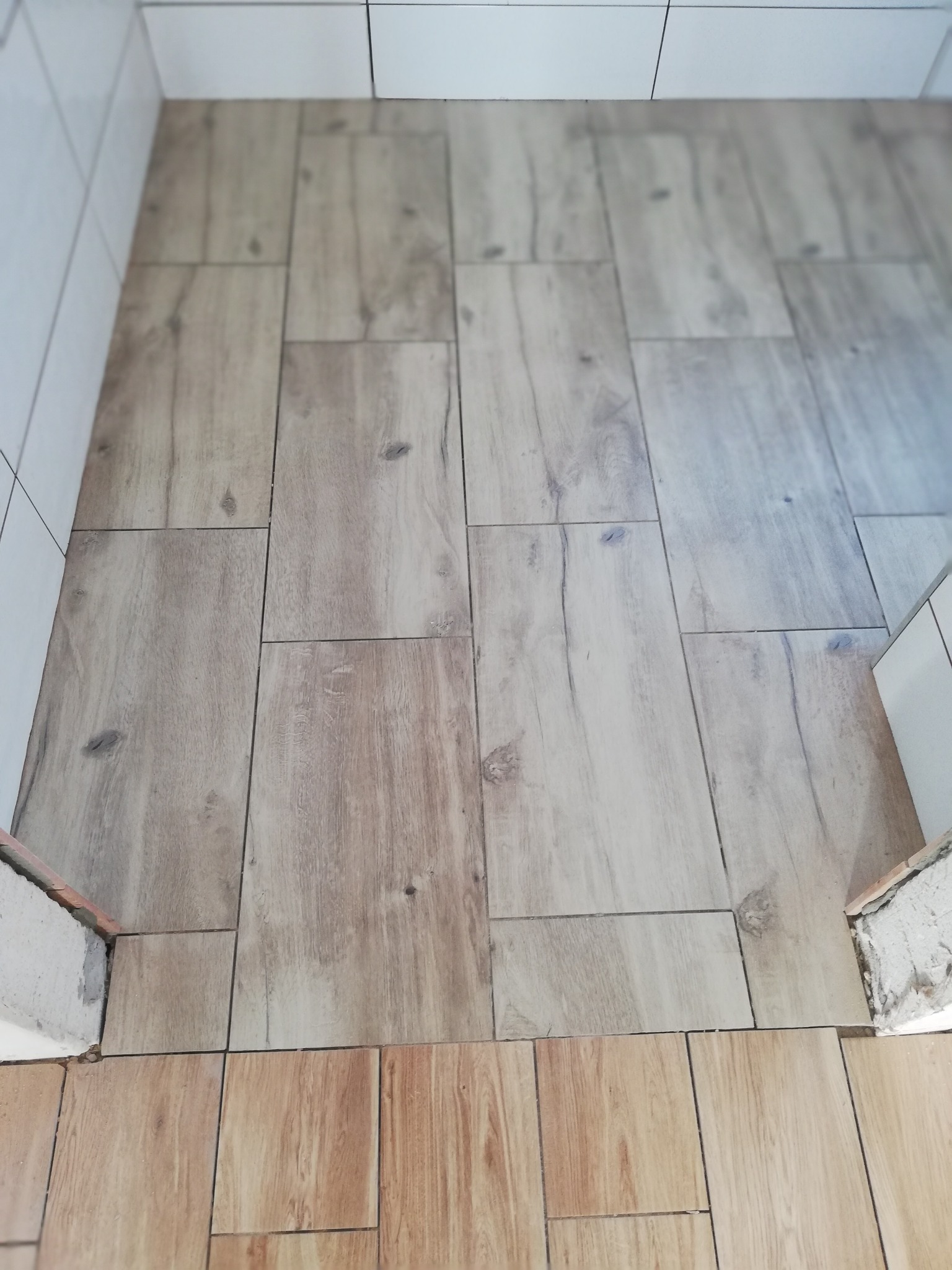 Ułożona podłoga w łazience z płytek imitujących drewno, widoczne fragmenty ścian z białymi płytkami i surowym wykończeniem przy podłodze.