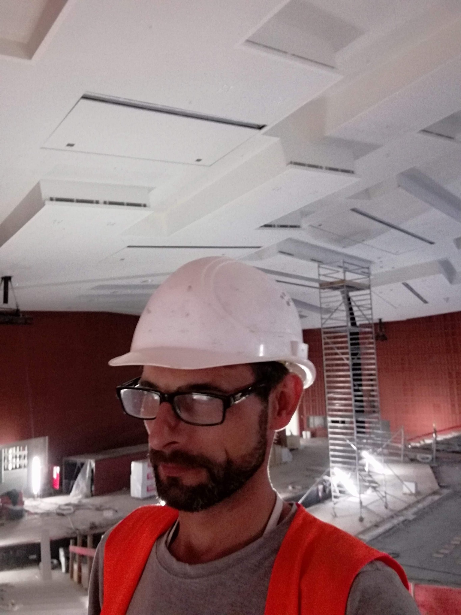 Pracownik w kasku i kamizelce odblaskowej na tle sufitu z płytami kartonowo-gipsowymi i rusztowania w trakcie prac wykończeniowych.