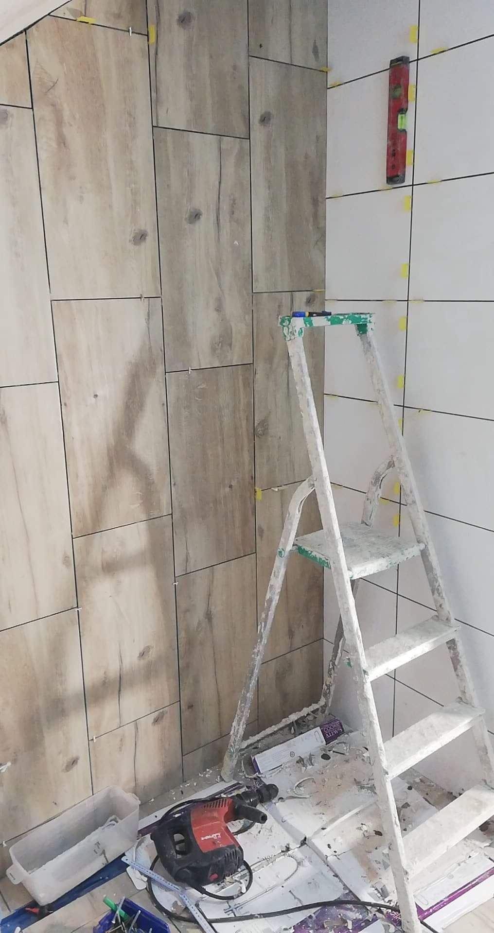 Fragment łazienki w trakcie układania płytek imitujących drewno na ścianie, widoczna drabina malarska, poziomica i wiertarka udarowa Hilti.