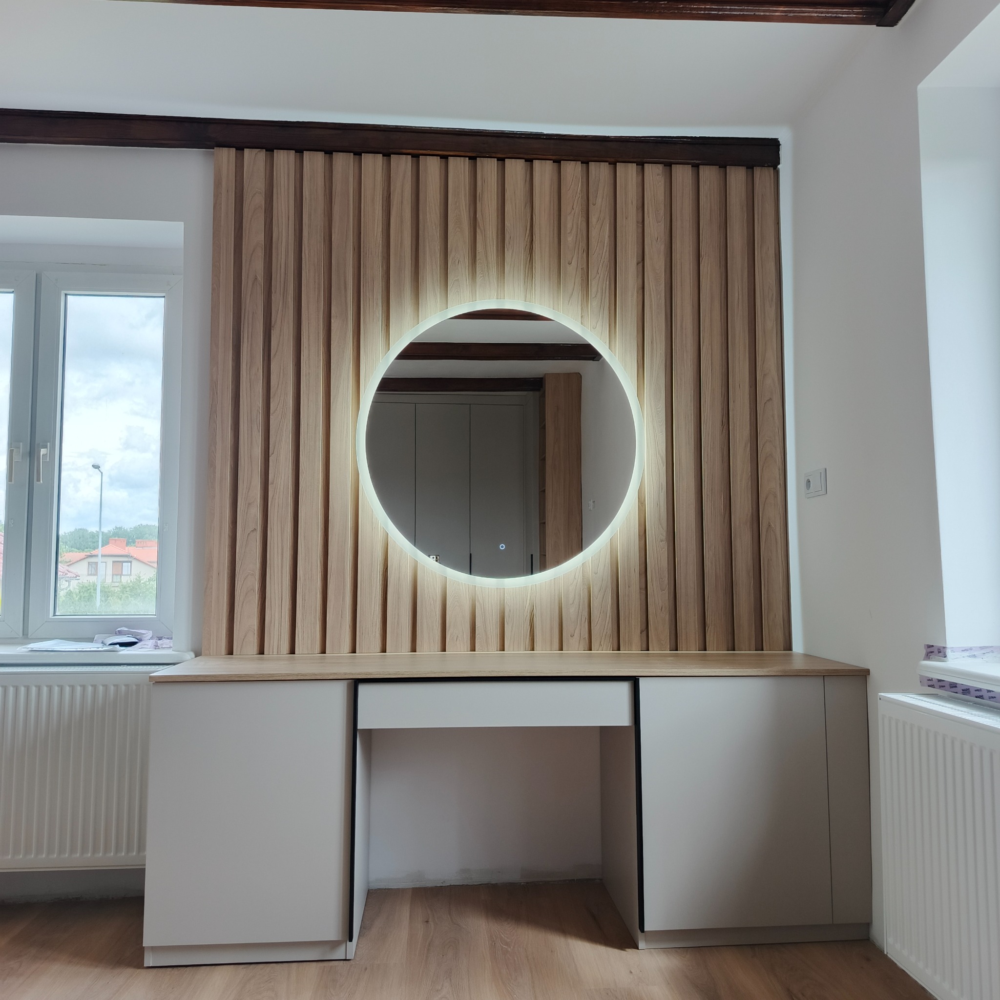 Nowoczesna toaletka z drewnianą lamelią i okrągłym lustrem z podświetleniem LED. Minimalistyczny design, białe szafki i drewniany blat. Widok na wnętrze przez lustro.
