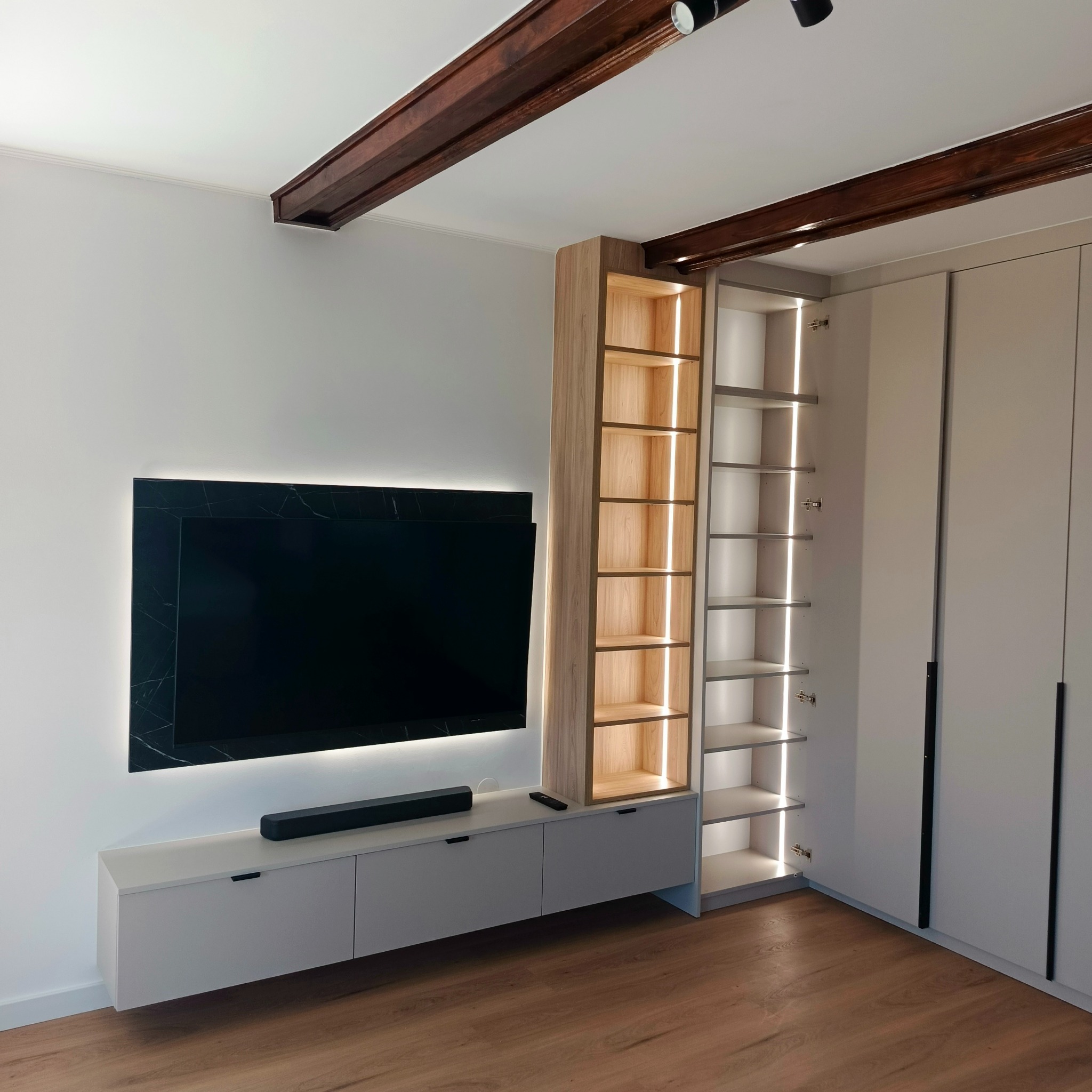 Nowoczesna zabudowa ściany z szafą, półkami i szafką RTV w odcieniach szarości i drewna. Podświetlenie LED dodaje wnętrzu elegancji. Widoczne drewniane belki stropowe.