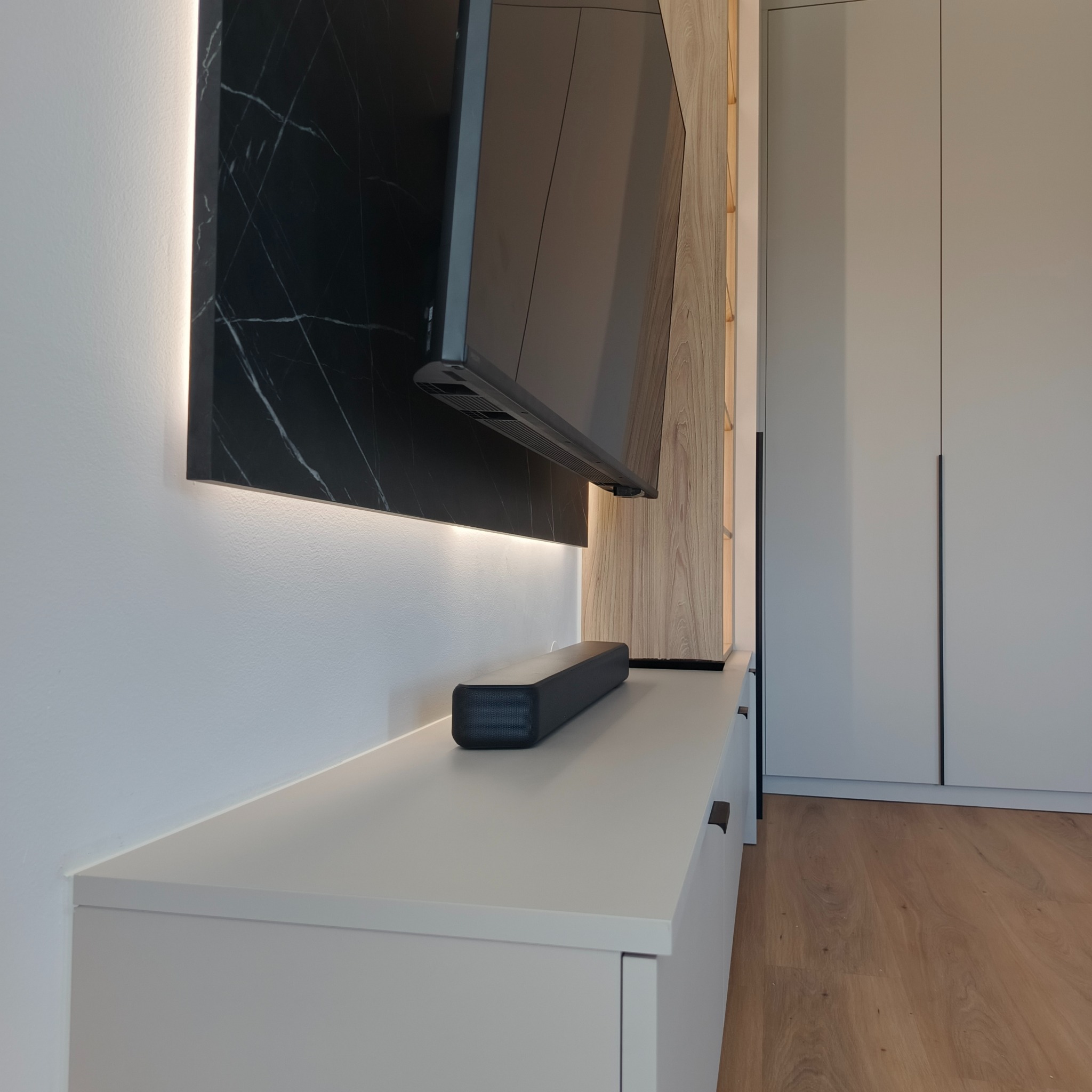 Nowoczesny salon z podświetlanym panelem z czarnego marmuru za telewizorem, szafka RTV z szufladami i drewniana zabudowa ściany. Minimalistyczny design wnętrza.