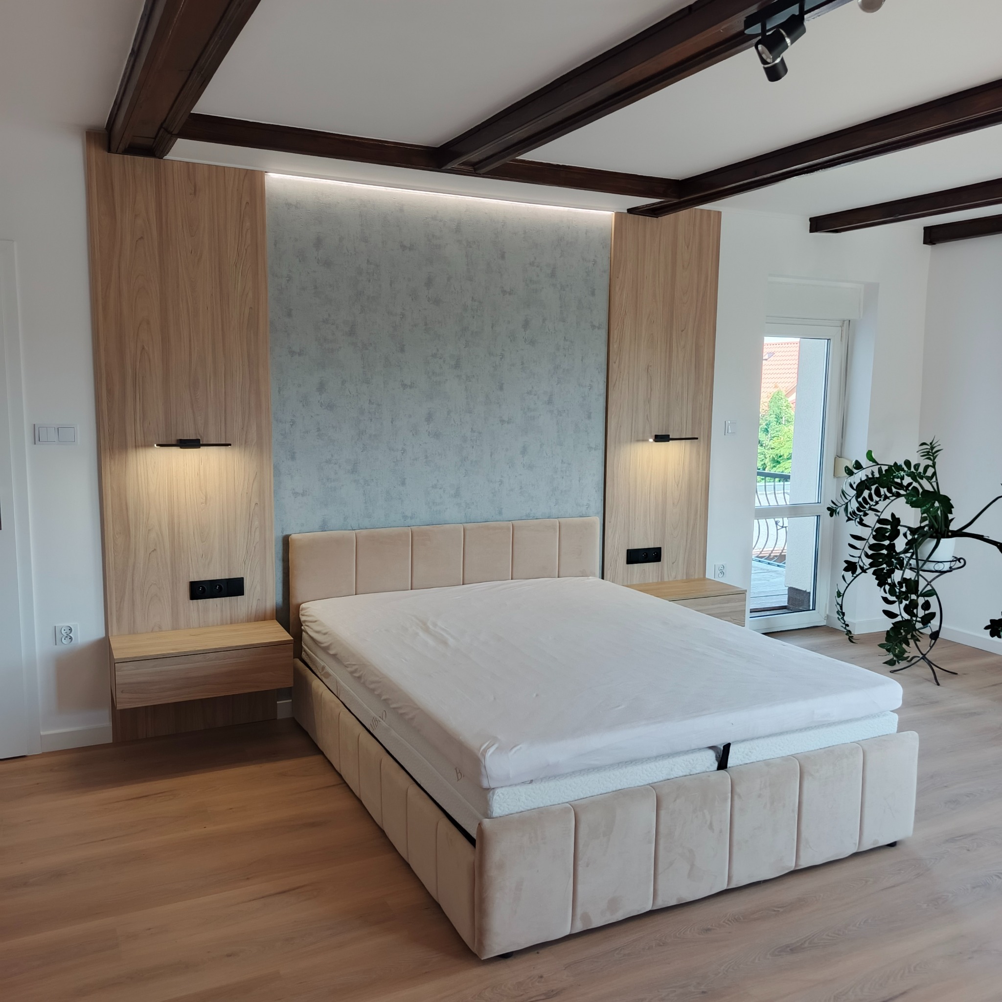 Nowoczesna sypialnia z beżowym łóżkiem, drewnianymi panelami i oświetleniem LED. Minimalistyczny design, drewniana podłoga i sufit z belkami.