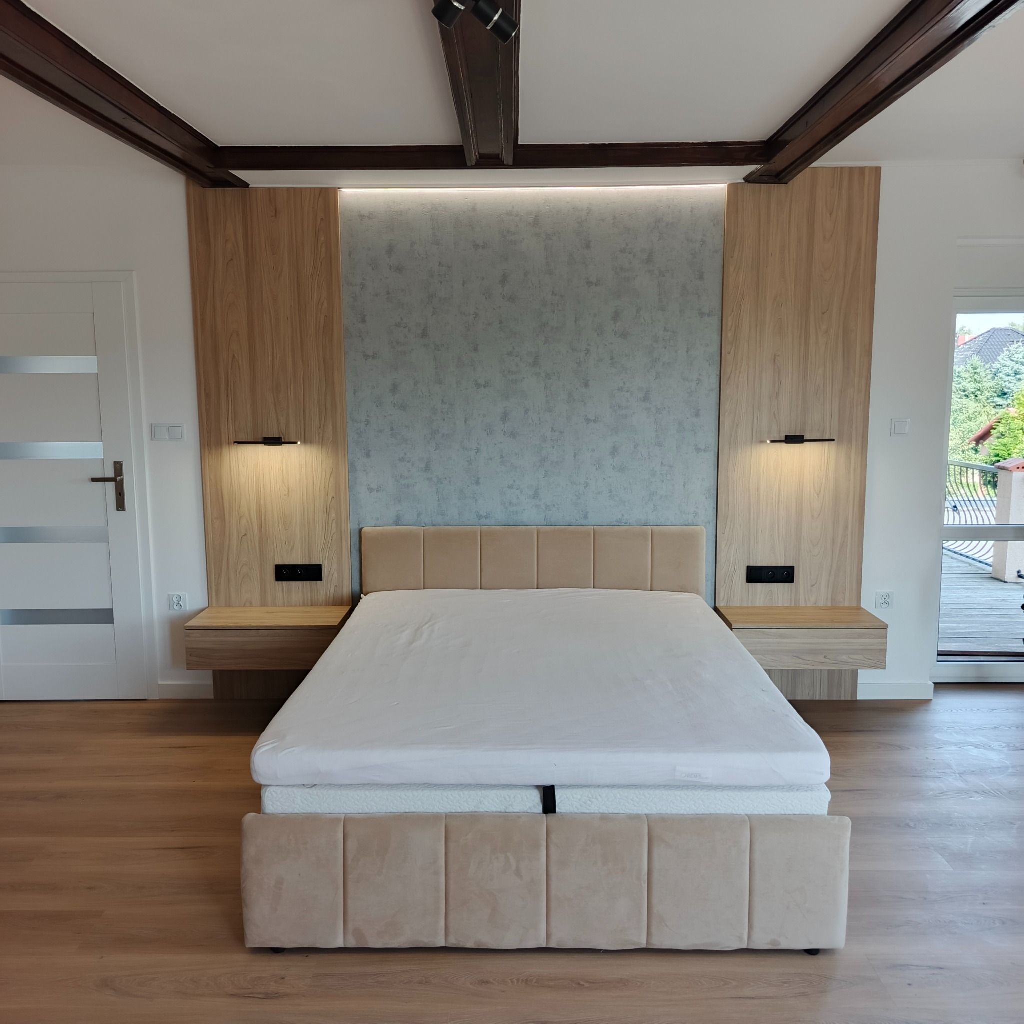 Nowoczesna sypialnia: drewniane panele ścienne z oświetleniem LED, beżowe tapicerowane łóżko z szafkami nocnymi i drewniana podłoga. Widoczne elementy dekoracyjne sufitu.
