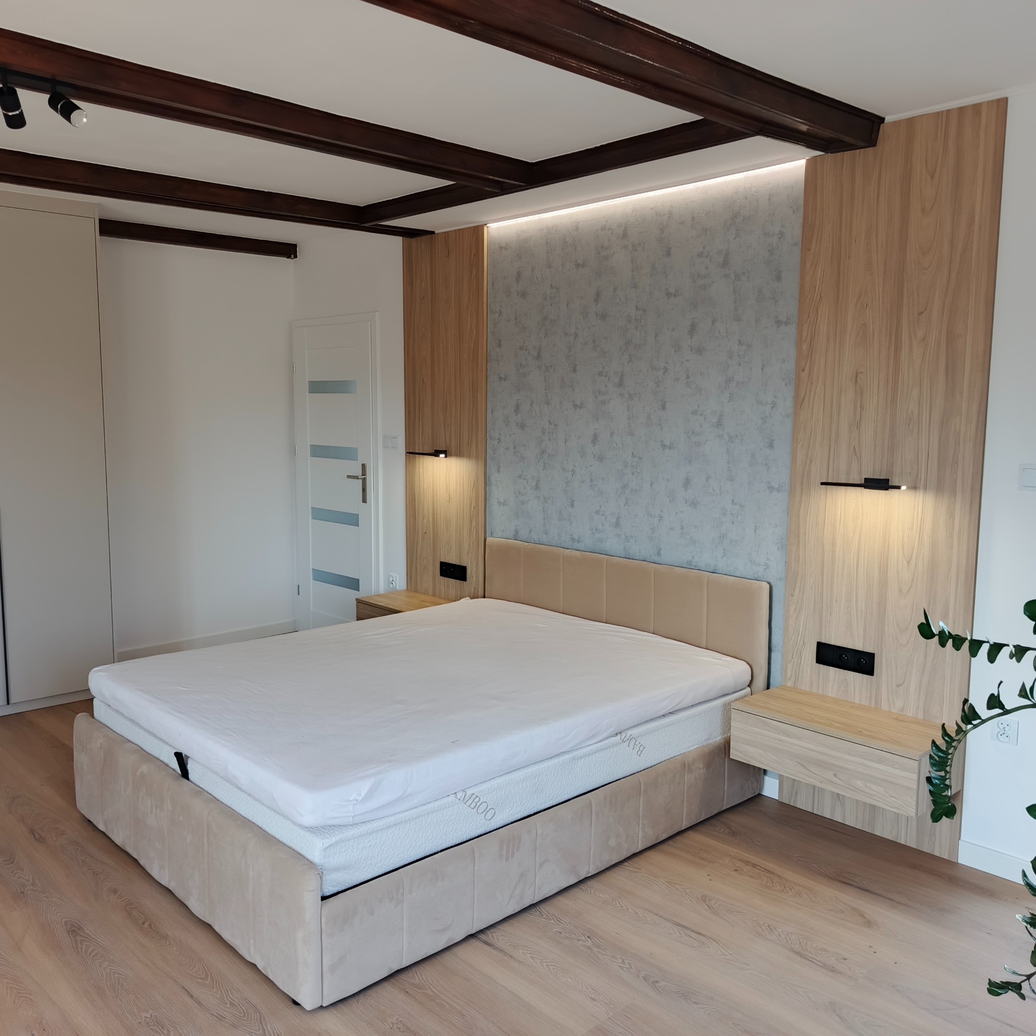Nowoczesna sypialnia z beżowym łóżkiem, drewnianą boazerią i nocnymi szafkami. Sufit z ciemnymi belkami. Minimalistyczny design, ciepłe oświetlenie.