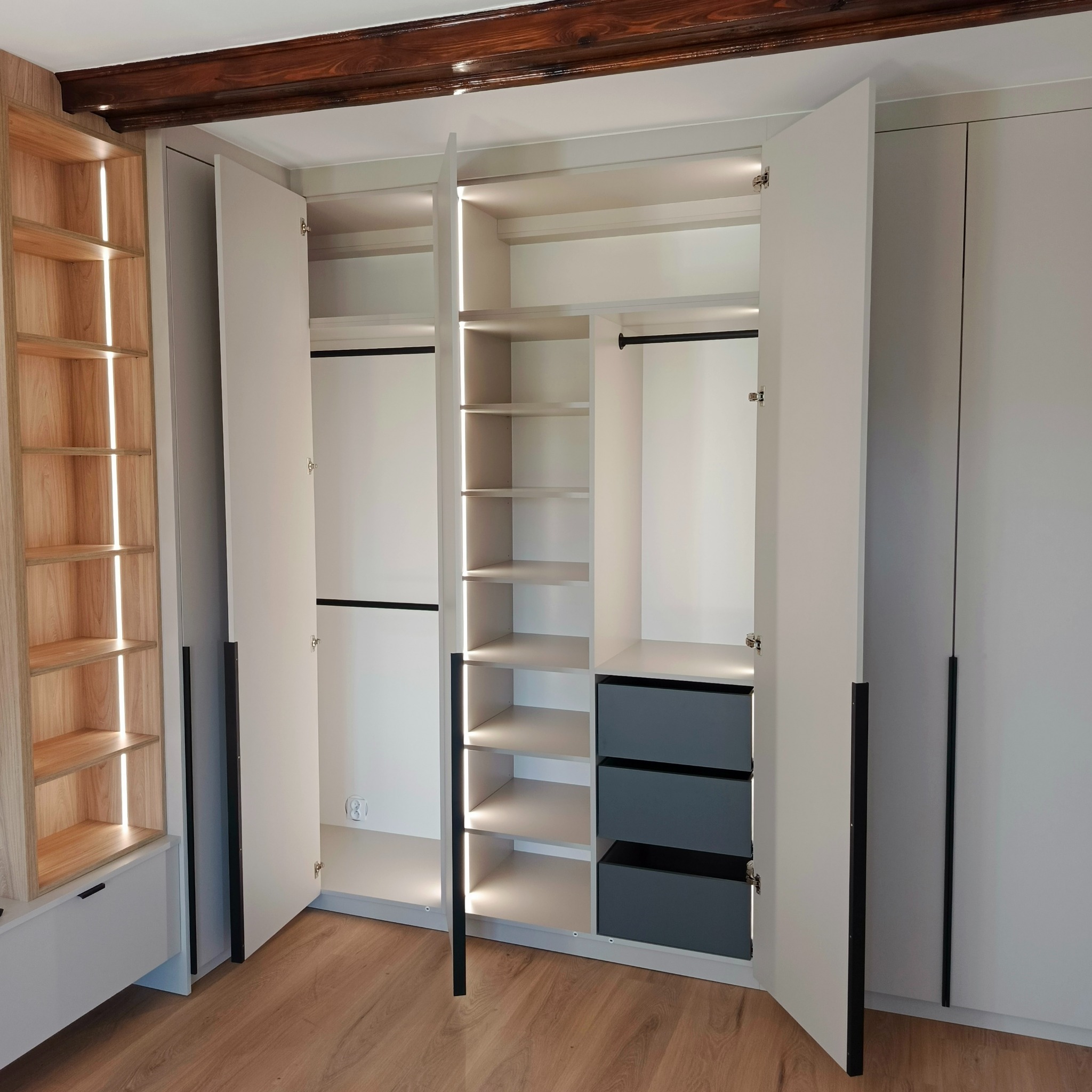 Otwarta szafa wnękowa z oświetleniem LED, widoczne półki, szuflady w odcieniach szarości i drewniane wykończenie. Na suficie widoczna drewniana belka.