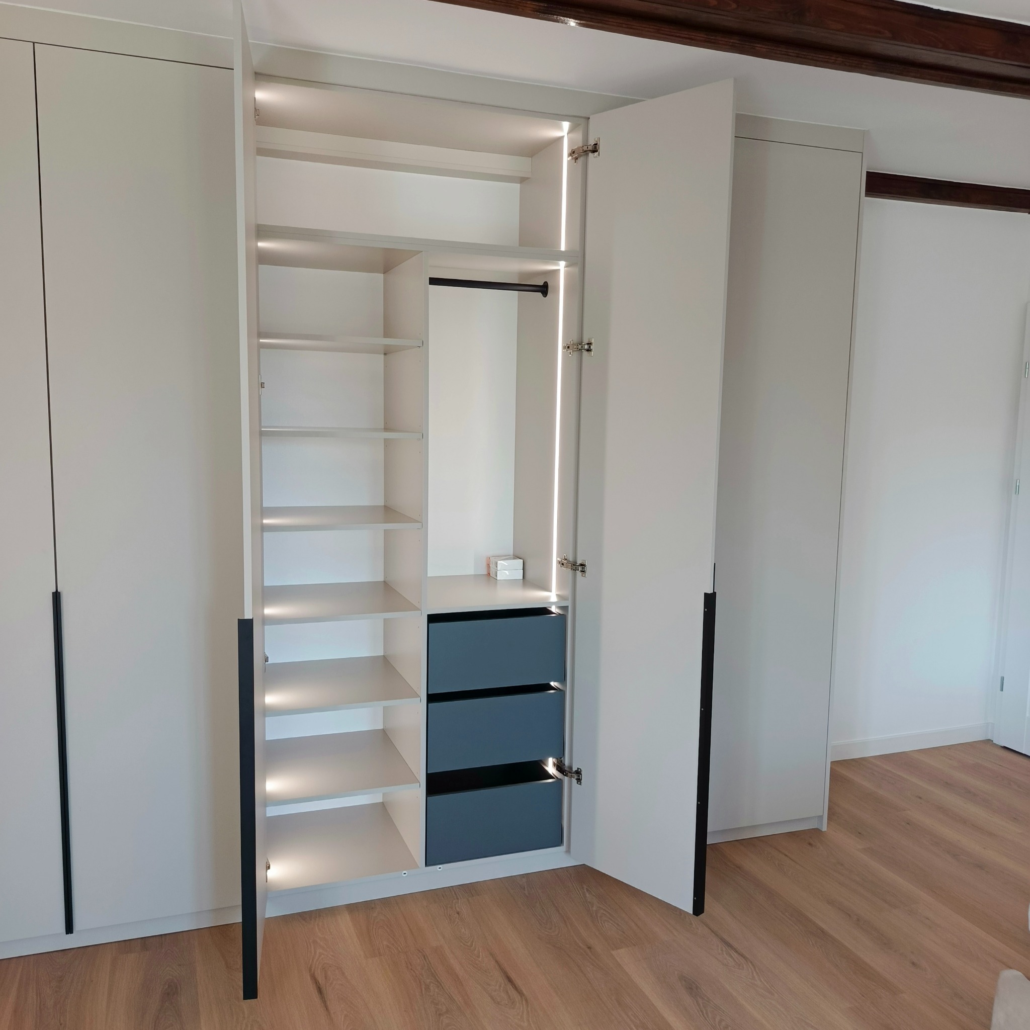 Nowoczesna szafa wnękowa z oświetleniem LED, otwarte drzwi ukazują półki i szuflady w odcieniach szarości. Widoczne drewniane belki stropowe.