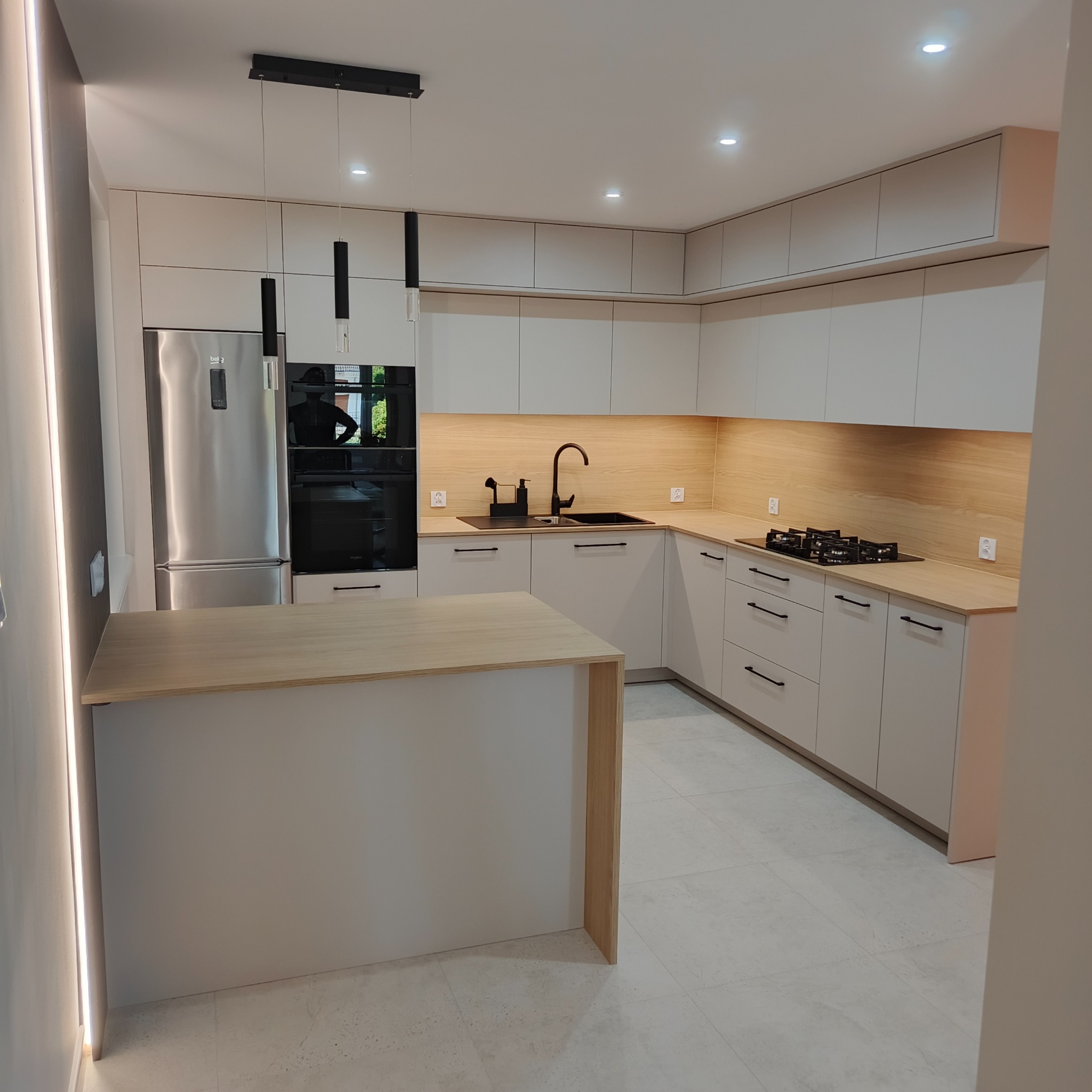 Jasna kuchnia z wyspą, minimalistyczne fronty, czarne akcenty armatury i oświetlenia. Blat z drewna, subtelne oświetlenie LED, nowoczesny design.