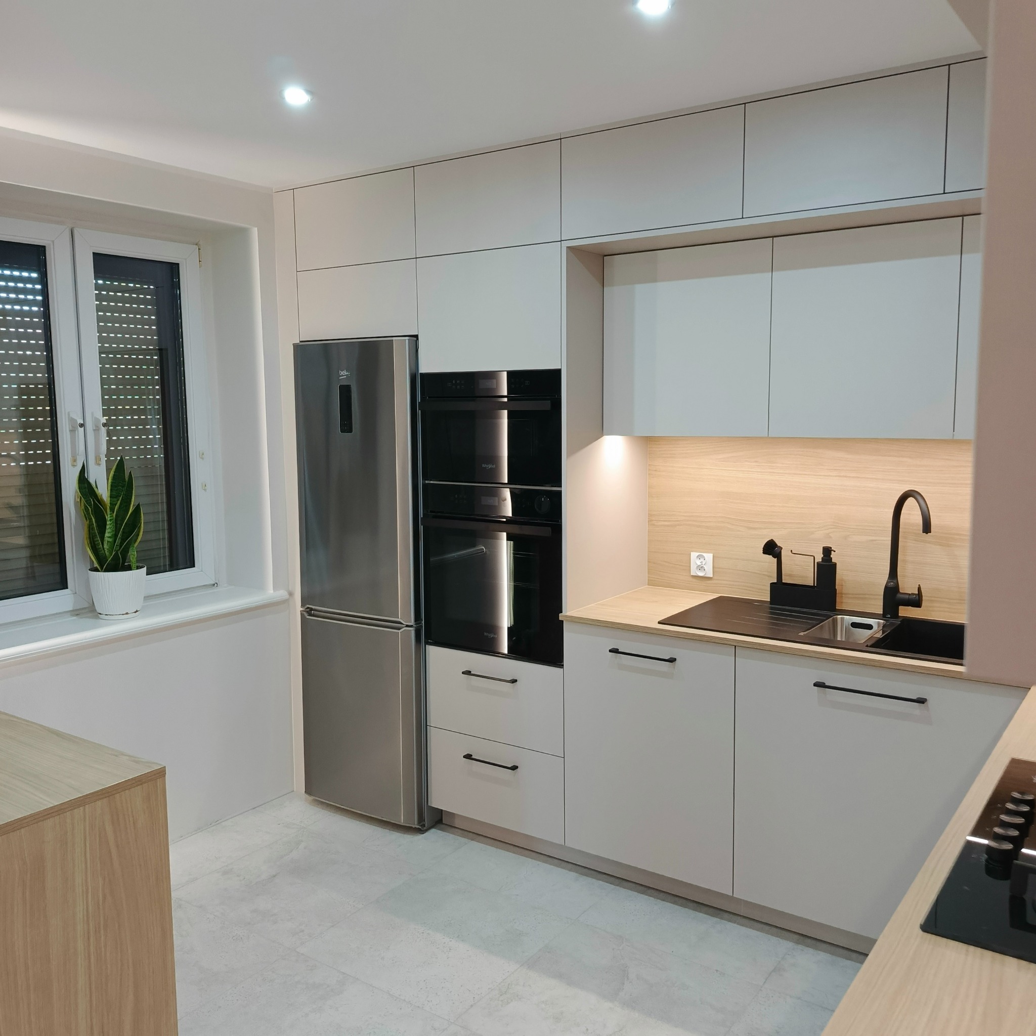 Nowoczesna kuchnia z białymi szafkami, drewnianym blatem i czarną armaturą. Na blacie stoi czarny zlew, obok okno z roletami i roślina doniczkowa.