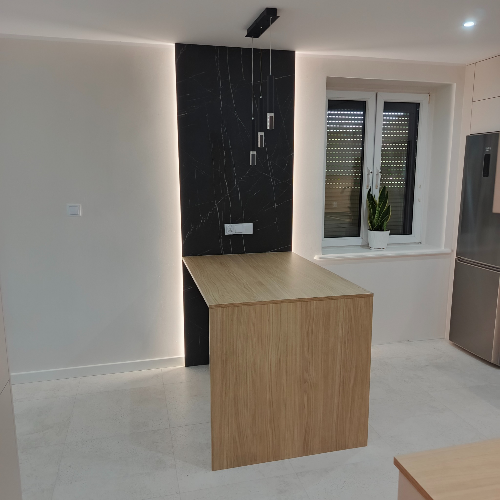 Nowoczesna kuchnia z drewnianym blatem i czarnym panelem ściennym z oświetleniem LED. Minimalistyczny design, roślina w oknie, widoczny fragment zabudowy kuchennej.