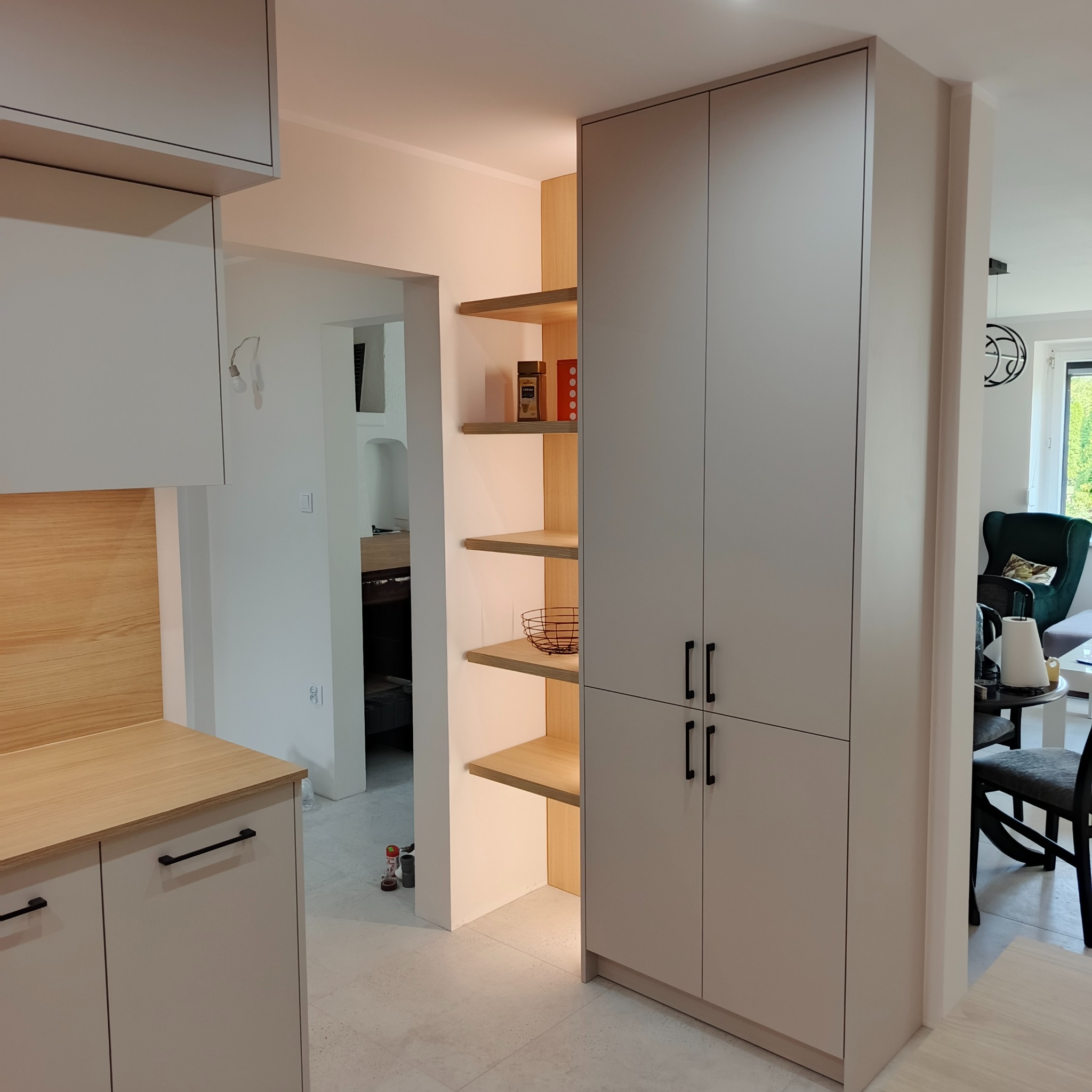 Jasne, nowoczesne meble kuchenne z drewnianymi akcentami i otwartymi półkami. Minimalistyczny design z czarnymi uchwytami. Widok na salon w tle.