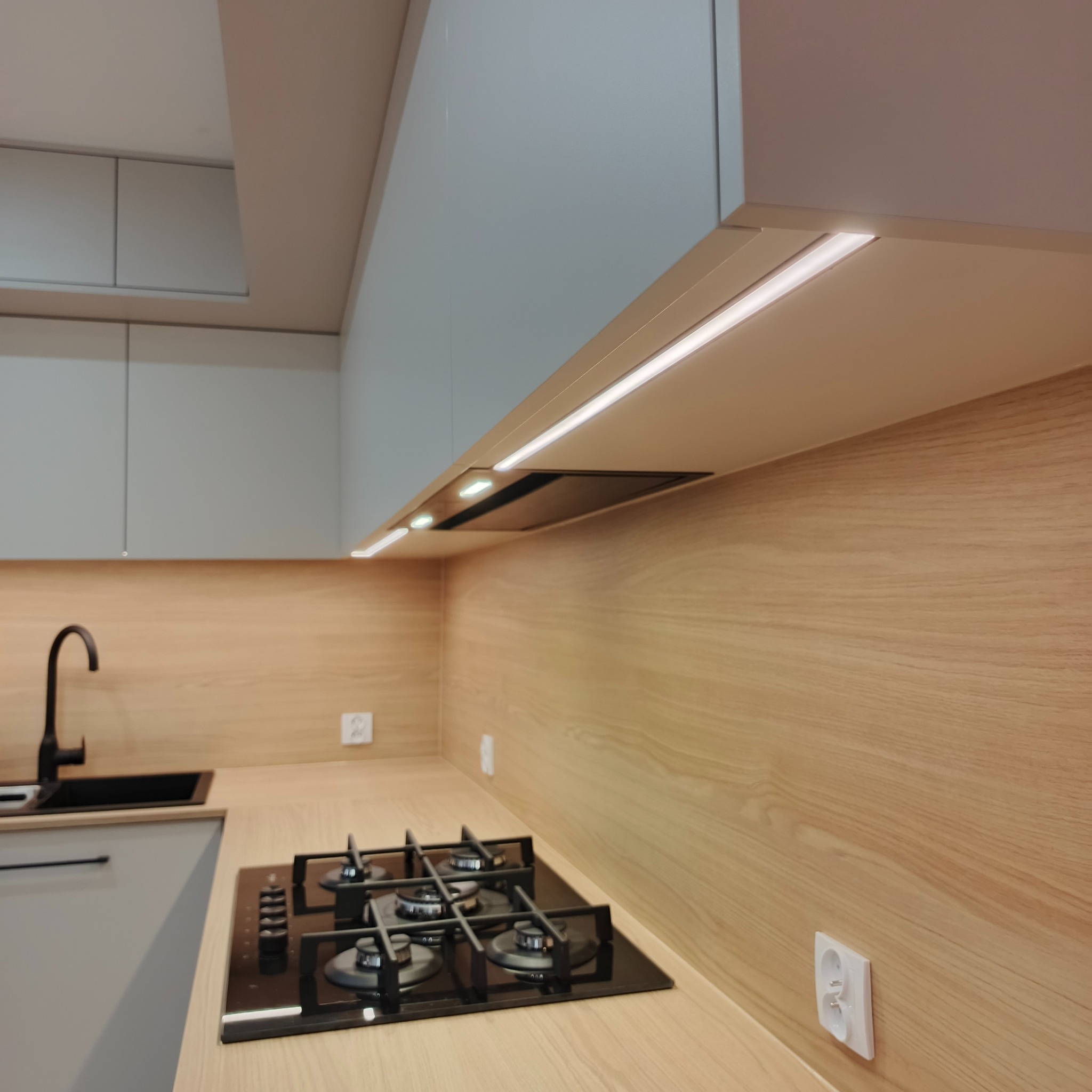 Jasna kuchnia z drewnianym blatem i płytą gazową. Oświetlenie LED pod szafkami. Minimalistyczny design z czarną armaturą i gładkimi frontami.