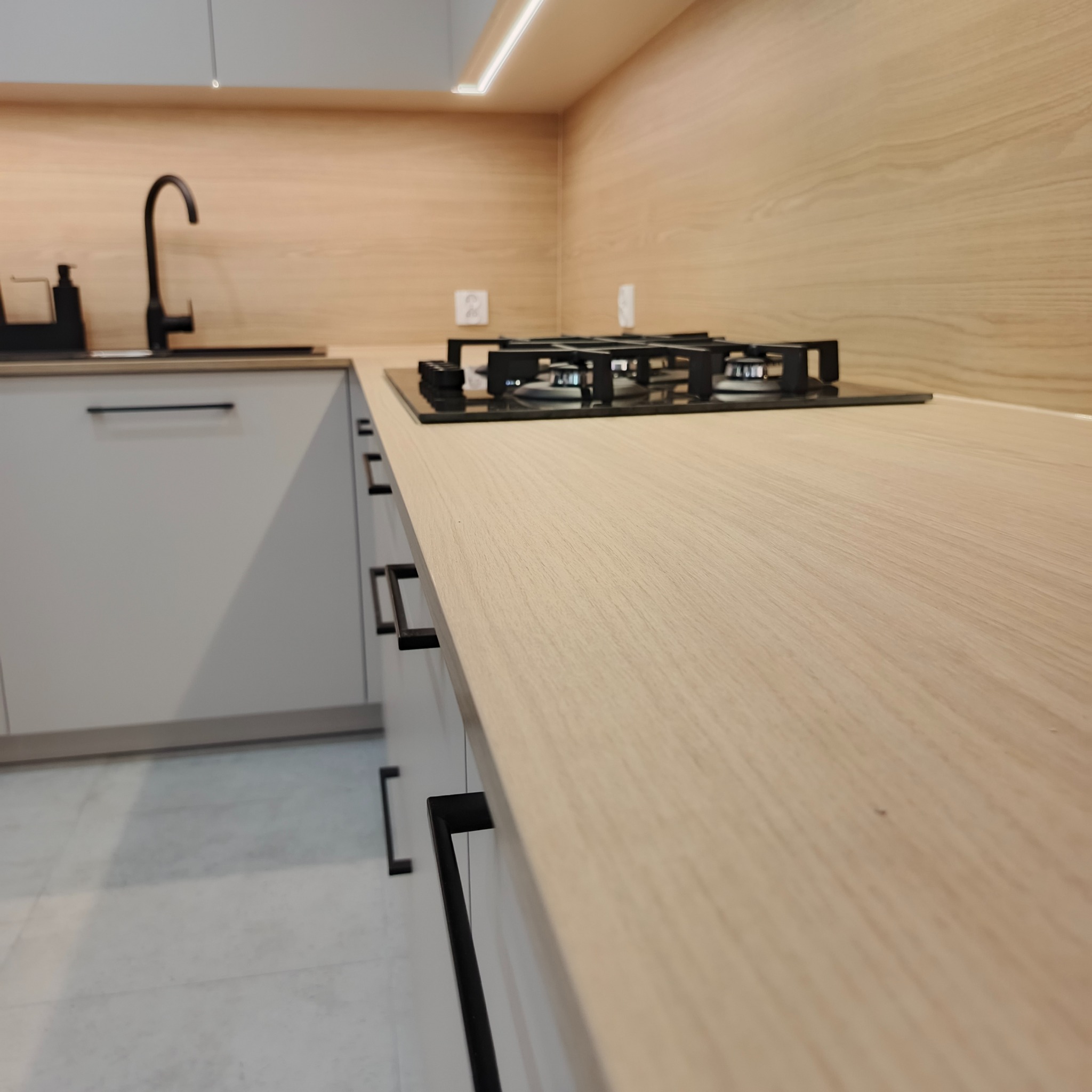 Jasny, drewniany blat kuchenny z czarną płytą gazową i minimalistycznymi uchwytami szafek. Ujęcie z niskiego kąta podkreśla fakturę blatu.