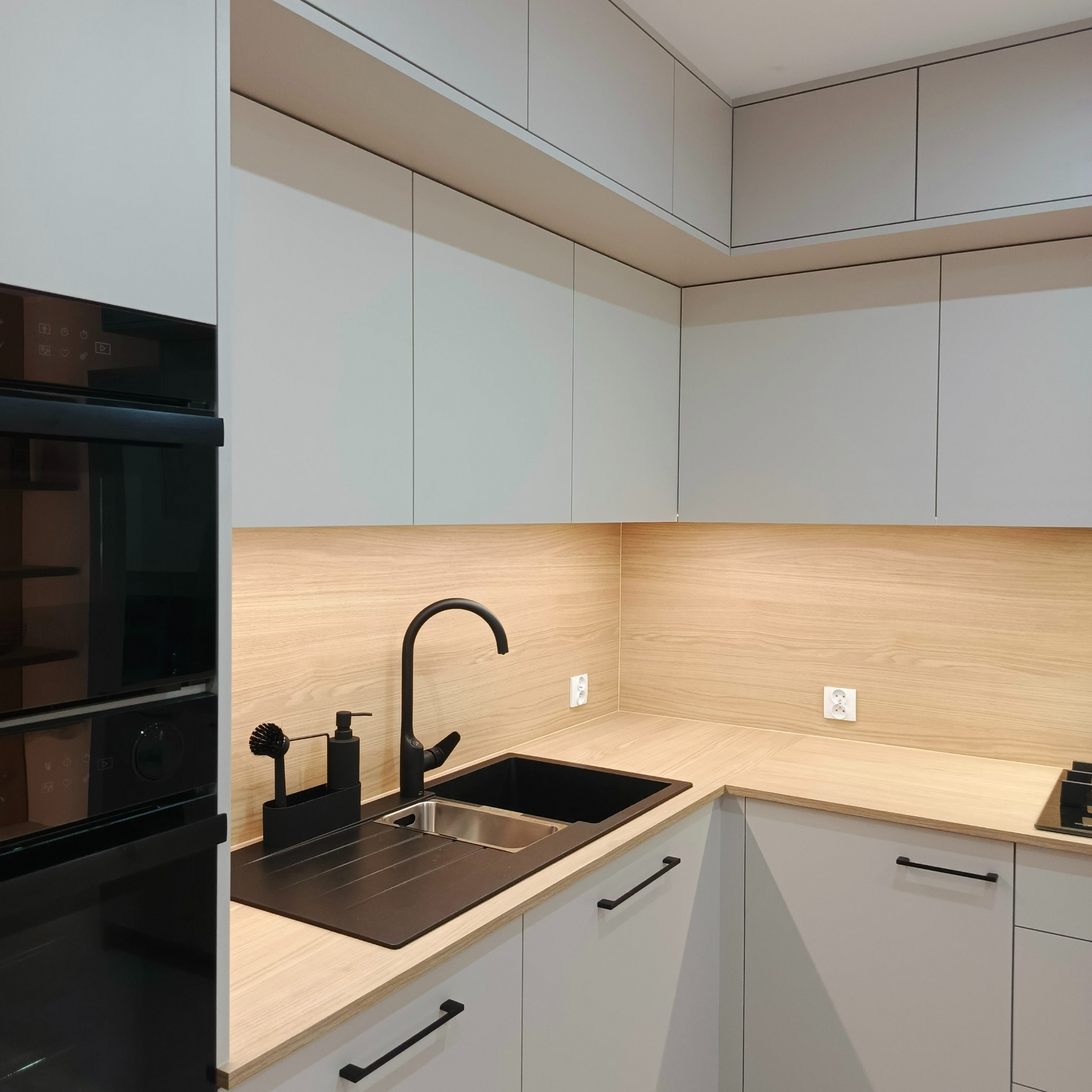 Nowoczesna kuchnia z drewnianym blatem, czarnym zlewem i armaturą, oraz szarymi szafkami. Widoczny fragment piekarnika i akcesoria kuchenne w minimalistycznym stylu.