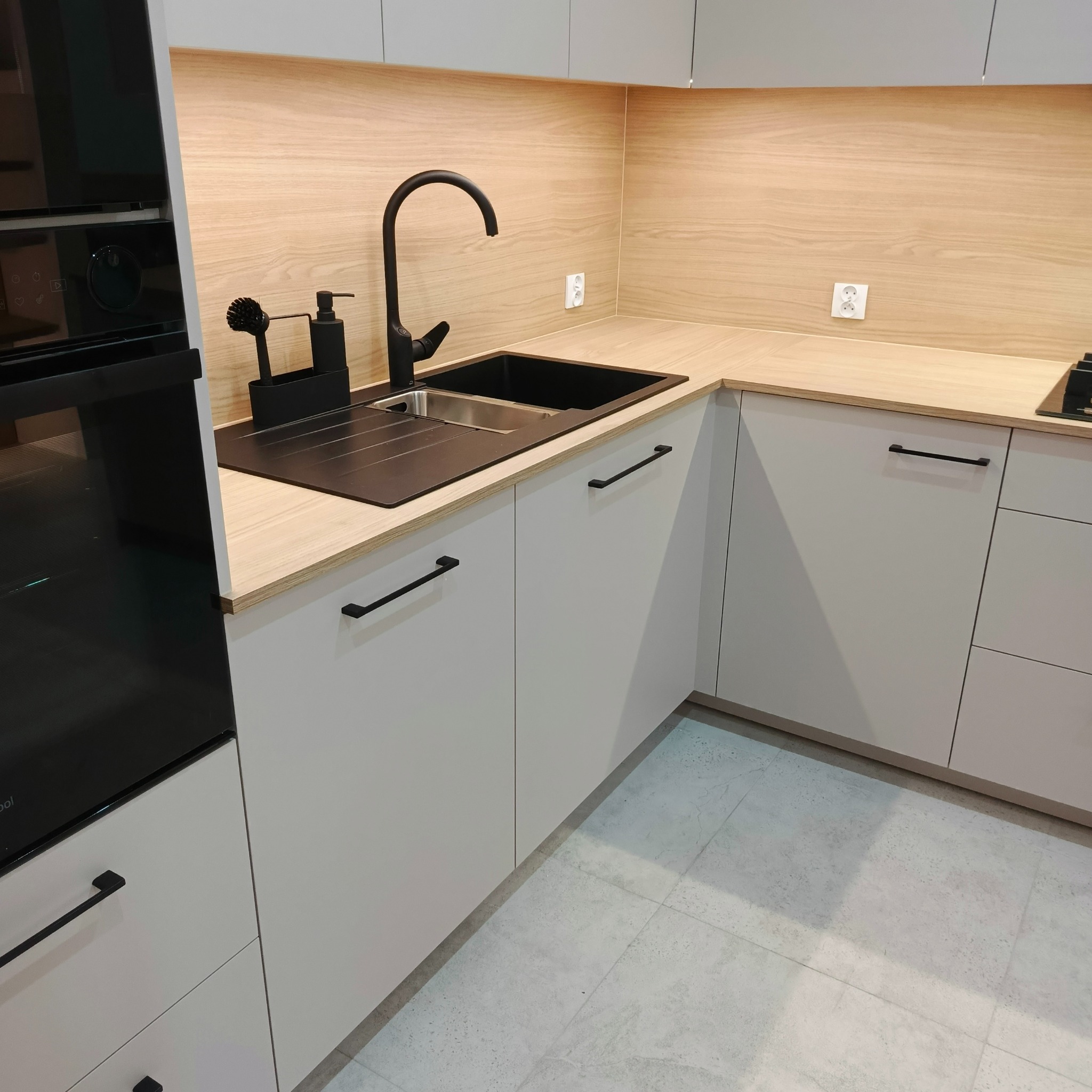 Nowoczesna kuchnia z szarymi szafkami, drewnianym blatem i czarną armaturą. Widoczny fragment piekarnika i akcesoria kuchenne w tonacji czerni.