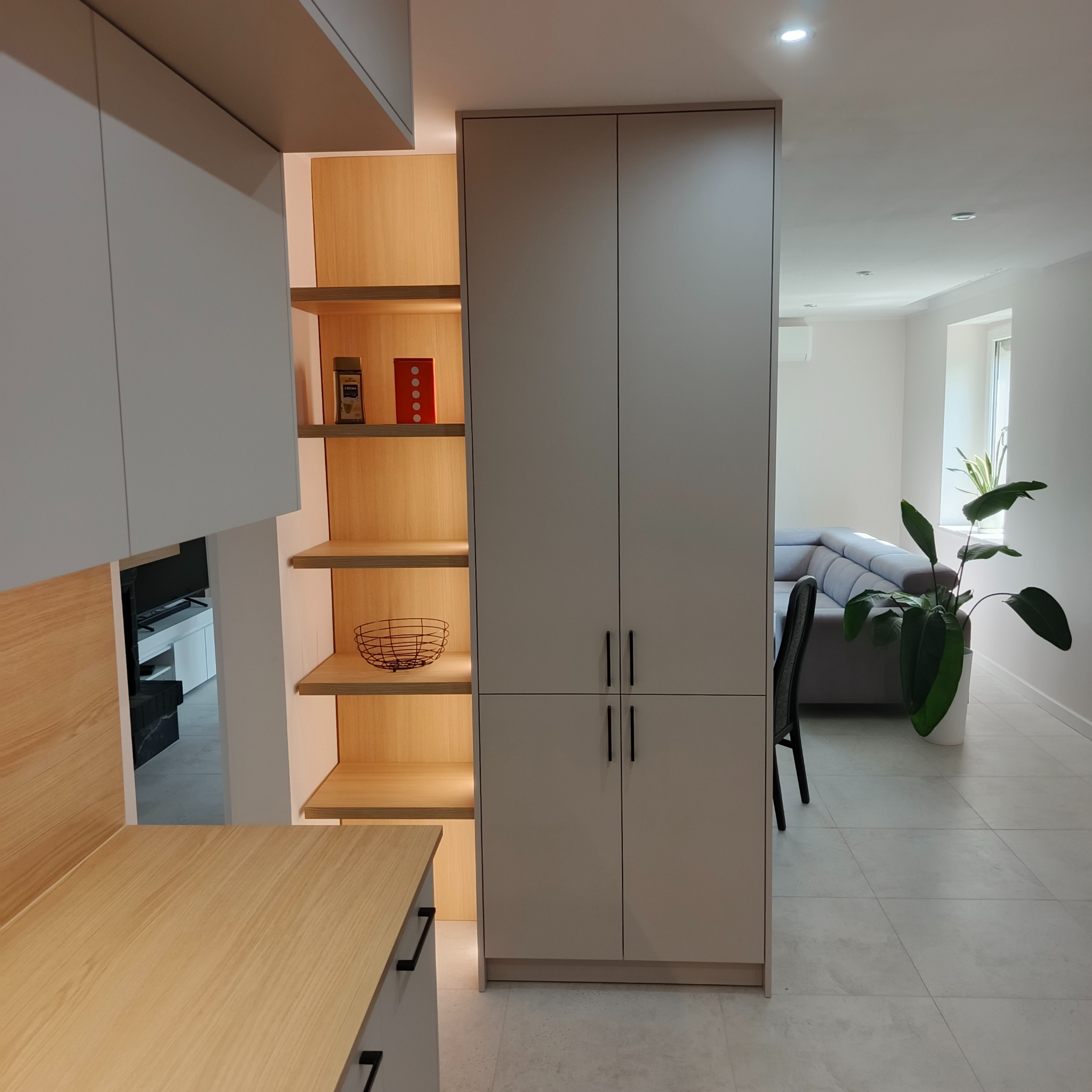 Nowoczesna zabudowa kuchenna w odcieniach szarości z drewnianymi akcentami i oświetleniem LED. Minimalistyczny design z otwartymi półkami i zamkniętymi szafkami.