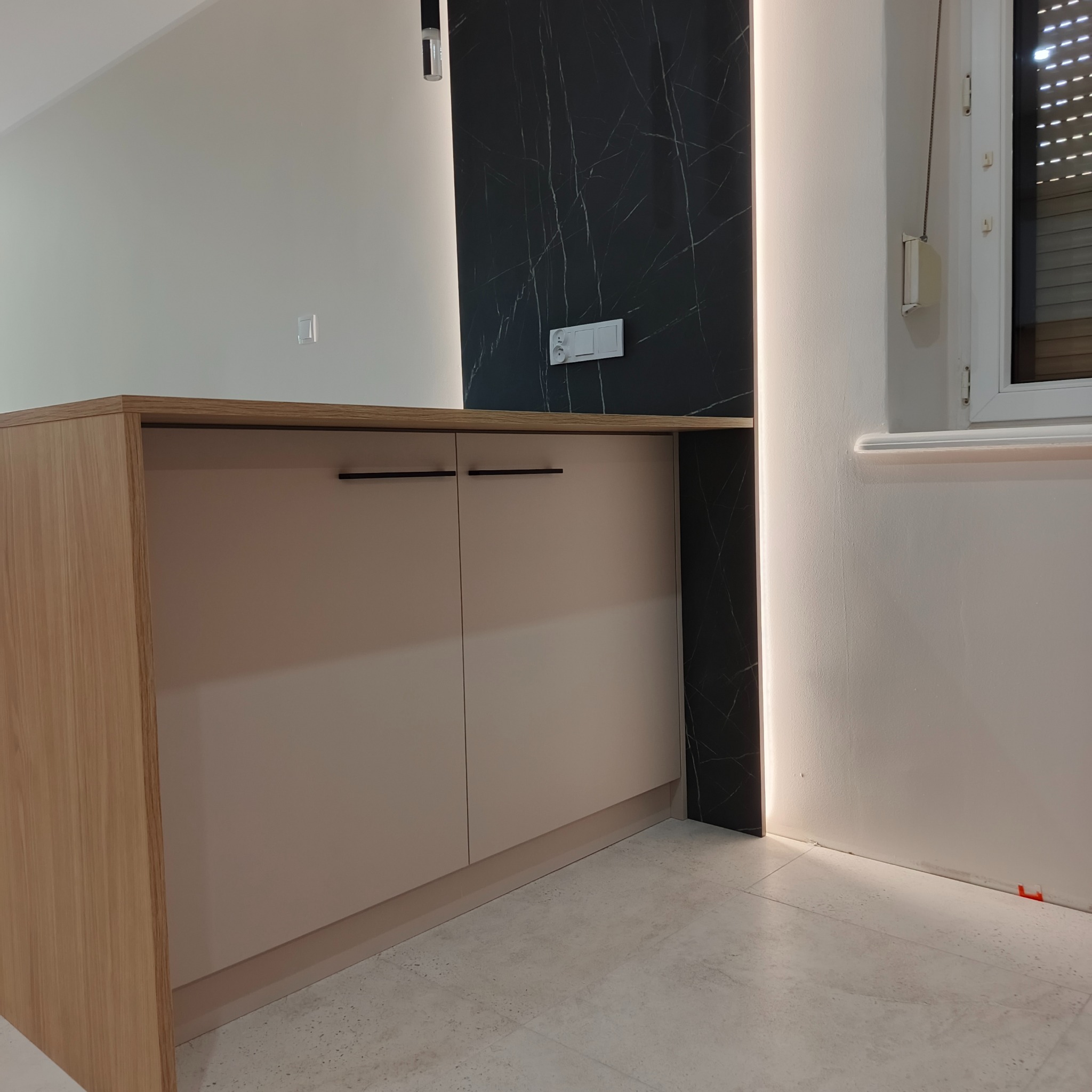 Nowoczesna zabudowa kuchenna z beżowymi frontami i drewnianym blatem, czarny panel ścienny z marmurowym wzorem, oświetlenie LED. Minimalistyczny design.
