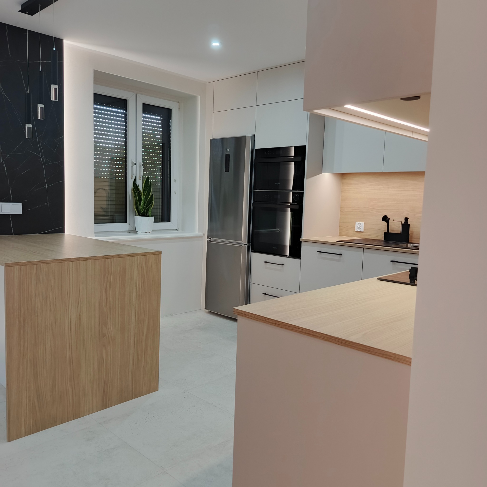 Nowoczesna kuchnia z wyspą: białe szafki, drewniane blaty, marmurowy akcent na ścianie, srebrna lodówka i czarne AGD. Minimalistyczny design, roślina w oknie.