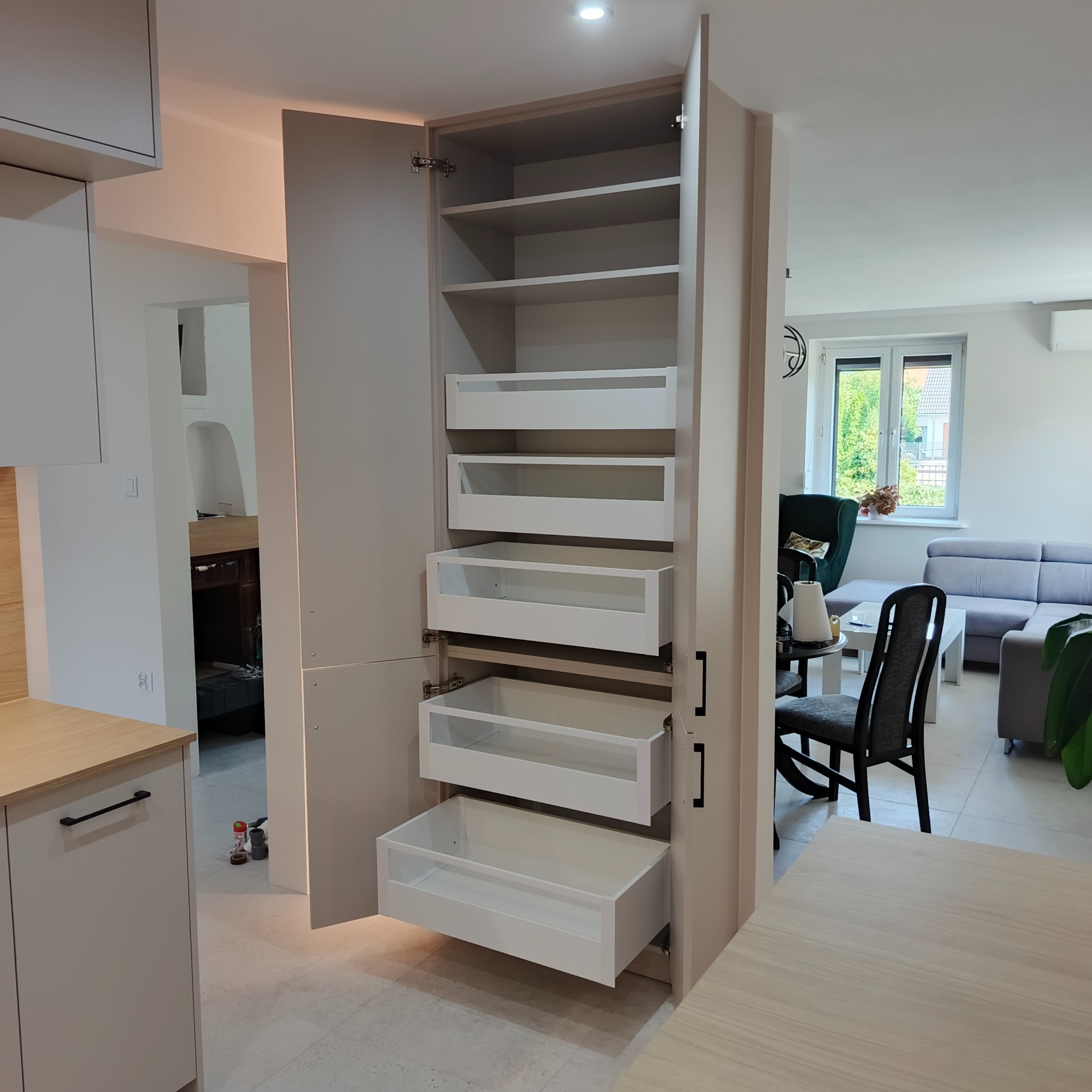 Szafa wnękowa z szufladami i półkami, jasne wykończenie, widok na salon w tle. Realizacja w Wilkowicach. Minimalistyczny design, czarne uchwyty, otwarte drzwi.