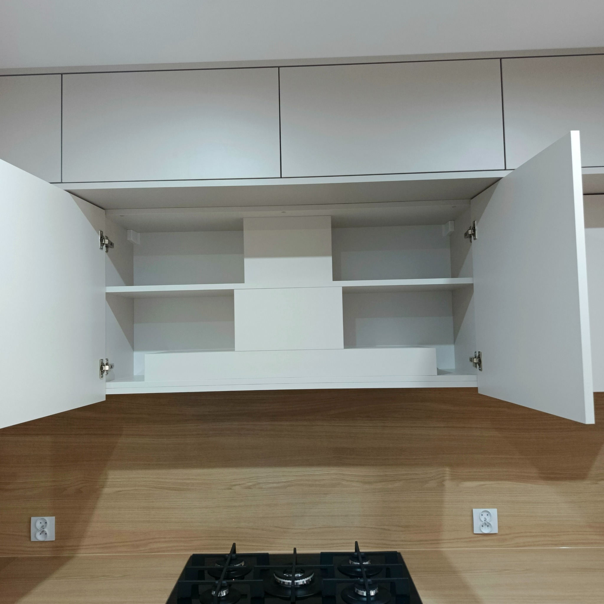 Białe szafki kuchenne nad blatem z drewnopodobnym wzorem, otwarte drzwiczki ukazują puste półki. Widoczna płyta grzewcza i gniazdka elektryczne w tle.