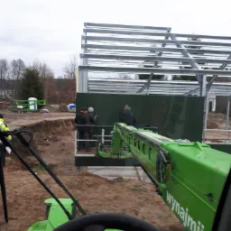 Widok z kabiny zielonej ładowarki teleskopowej na plac budowy hali z widoczną stalową konstrukcją szkieletową, montaż paneli ściennych przez pracowników na podnośniku i teren z wykopem.