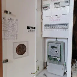 Otwarta skrzynka elektryczna z widocznym licznikiem, bezpiecznikami, wyłącznikami i listą obwodów, klucze leżące na dnie obudowy, ściana w tle z widocznymi uszkodzeniami tynku.