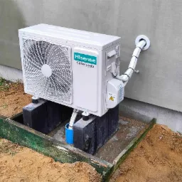 Zewnętrzna jednostka klimatyzatora Hisense zamontowana na betonowych blokach, z widocznymi przewodami wchodzącymi do budynku przez otwór w ścianie, obok widoczny fragment skrzyni.