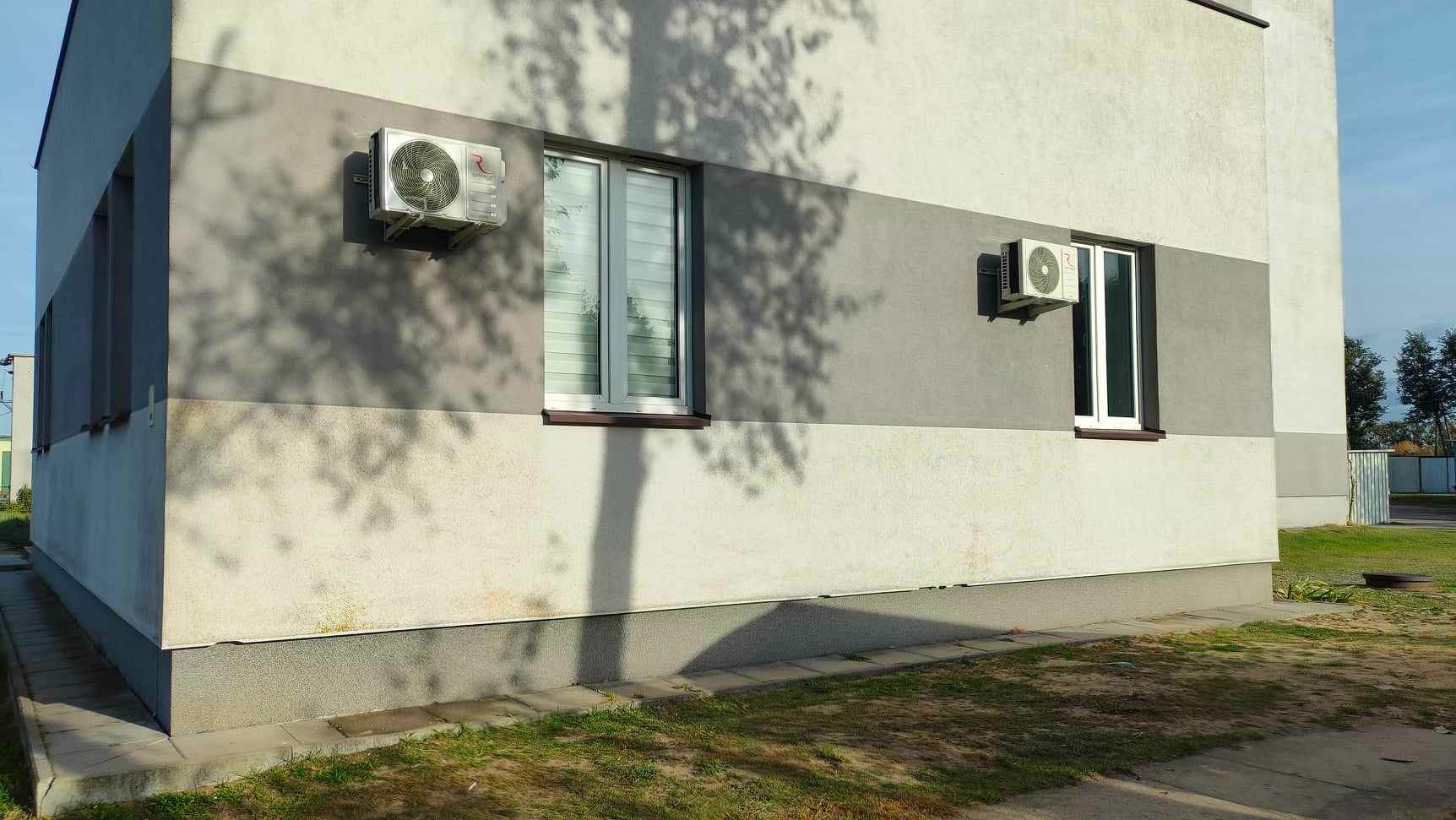 Dwa klimatyzatory zamontowane na zewnętrznej ścianie budynku biurowego o dwukolorowej elewacji, z widocznymi oknami i cieniem drzewa.
