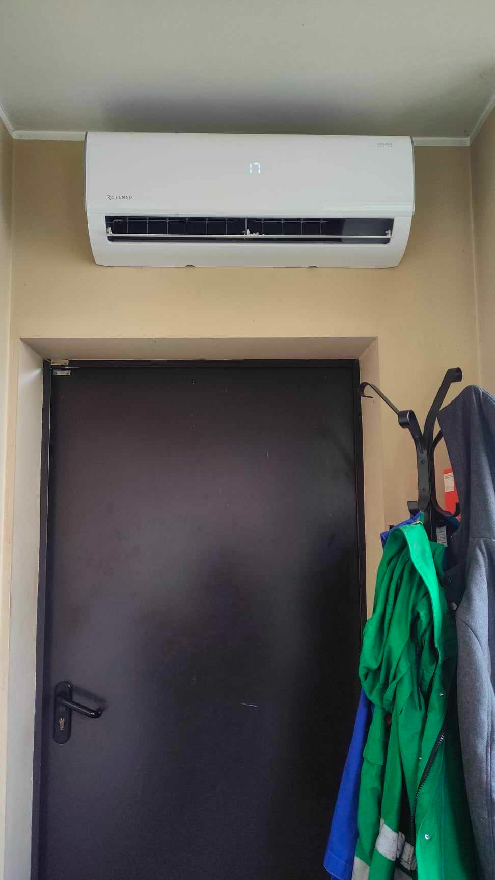 Biały klimatyzator Rotenso na ścianie nad czarnymi drzwiami, obok wieszak z ubraniami.