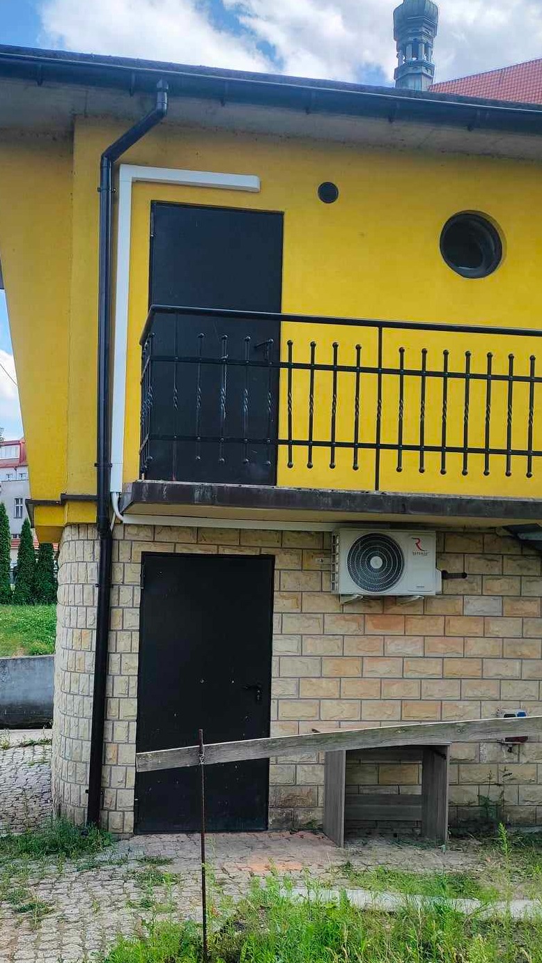 Żółty budynek z zewnętrzną jednostką klimatyzacyjną zamontowaną na ceglanej ścianie, czarne drzwi i balkon z balustradą.