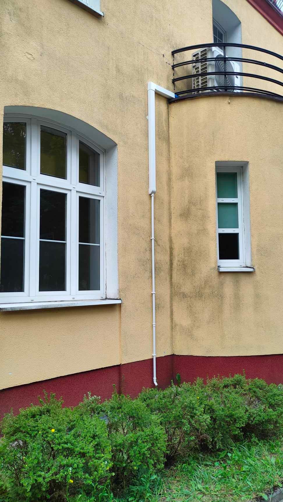 Żółta elewacja budynku z dwoma oknami, balkonem i zewnętrzną jednostką klimatyzacji z odprowadzeniem skroplin w białej rurze PCV.