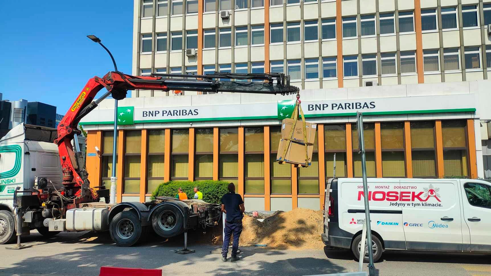 Rozładunek klimatyzatorów GREE z samochodu ciężarowego z dźwigiem Palfinger przed budynkiem BNP Paribas, widoczny samochód serwisowy firmy Nosek z logo Midea, Daikin i Mitsubishi Electric.