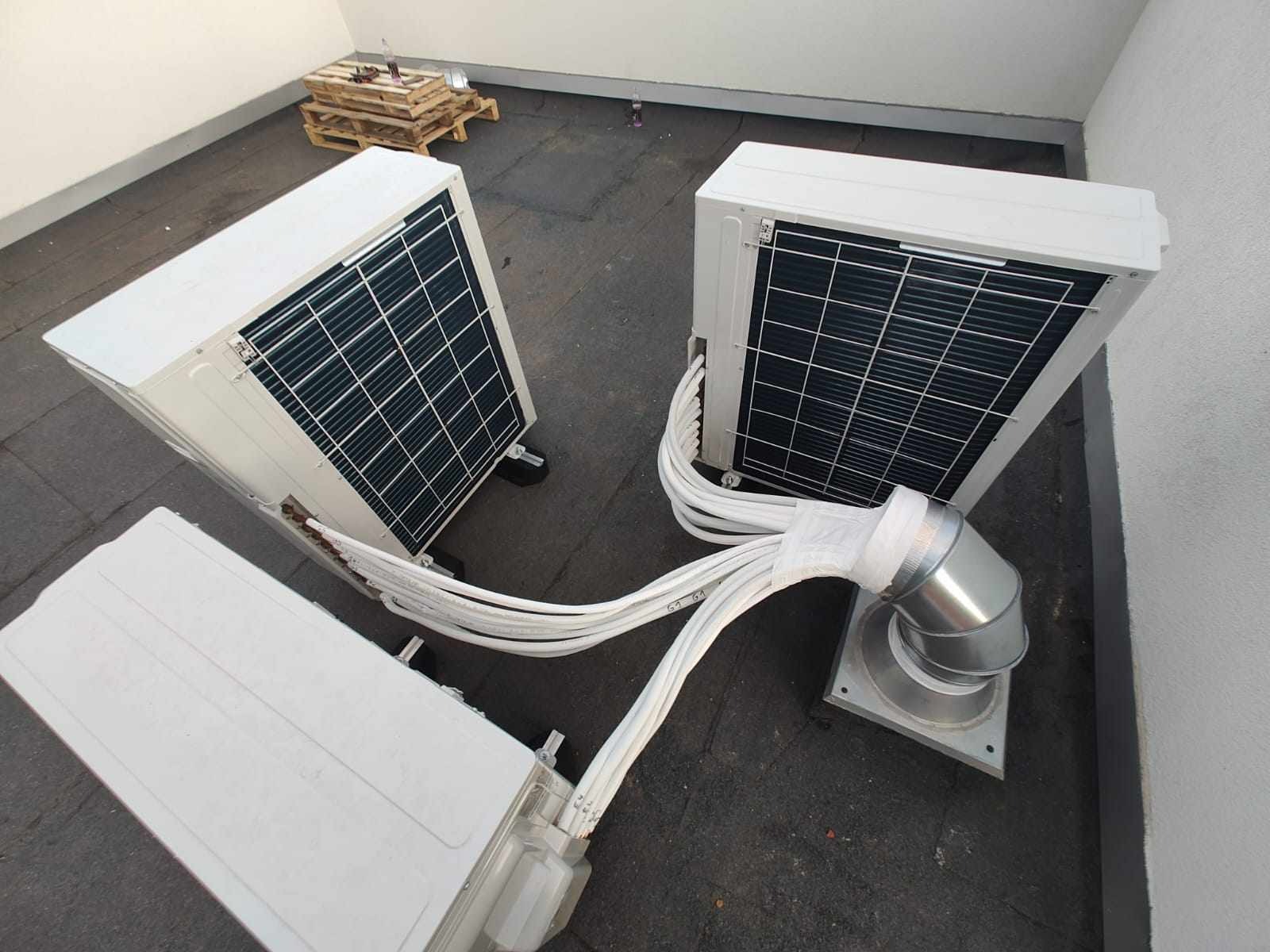 Trzy zewnętrzne jednostki klimatyzacyjne na płaskim dachu, połączone rurami, widoczne palety w tle.