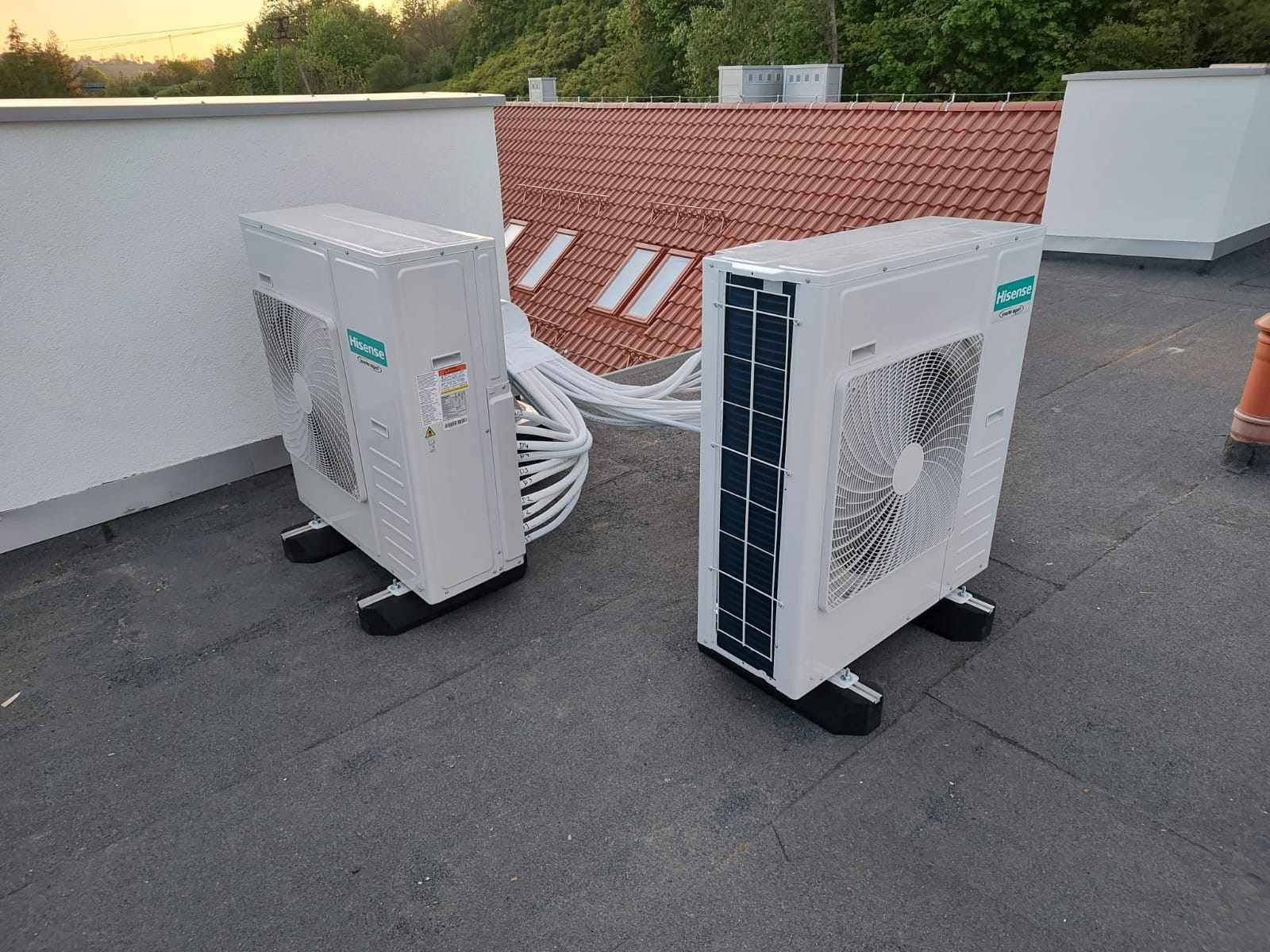 Dwa białe agregaty klimatyzacyjne marki Hisense, zamontowane na płaskim dachu budynku z widocznymi przewodami i fragmentem dachu skośnego z oknami.