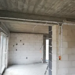 Surowy stan wnętrza z widocznymi przewodami elektrycznymi ułożonymi w korytkach na ścianach i suficie, widoczna puszka elektryczna na ścianie z betonu komórkowego.