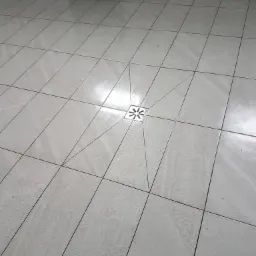 Jasnoszare płytki podłogowe ułożone pod kątem, z odpływem liniowym w kształcie kwiatu w centralnej części pomieszczenia, widoczny grzejnik na ścianie w tle.