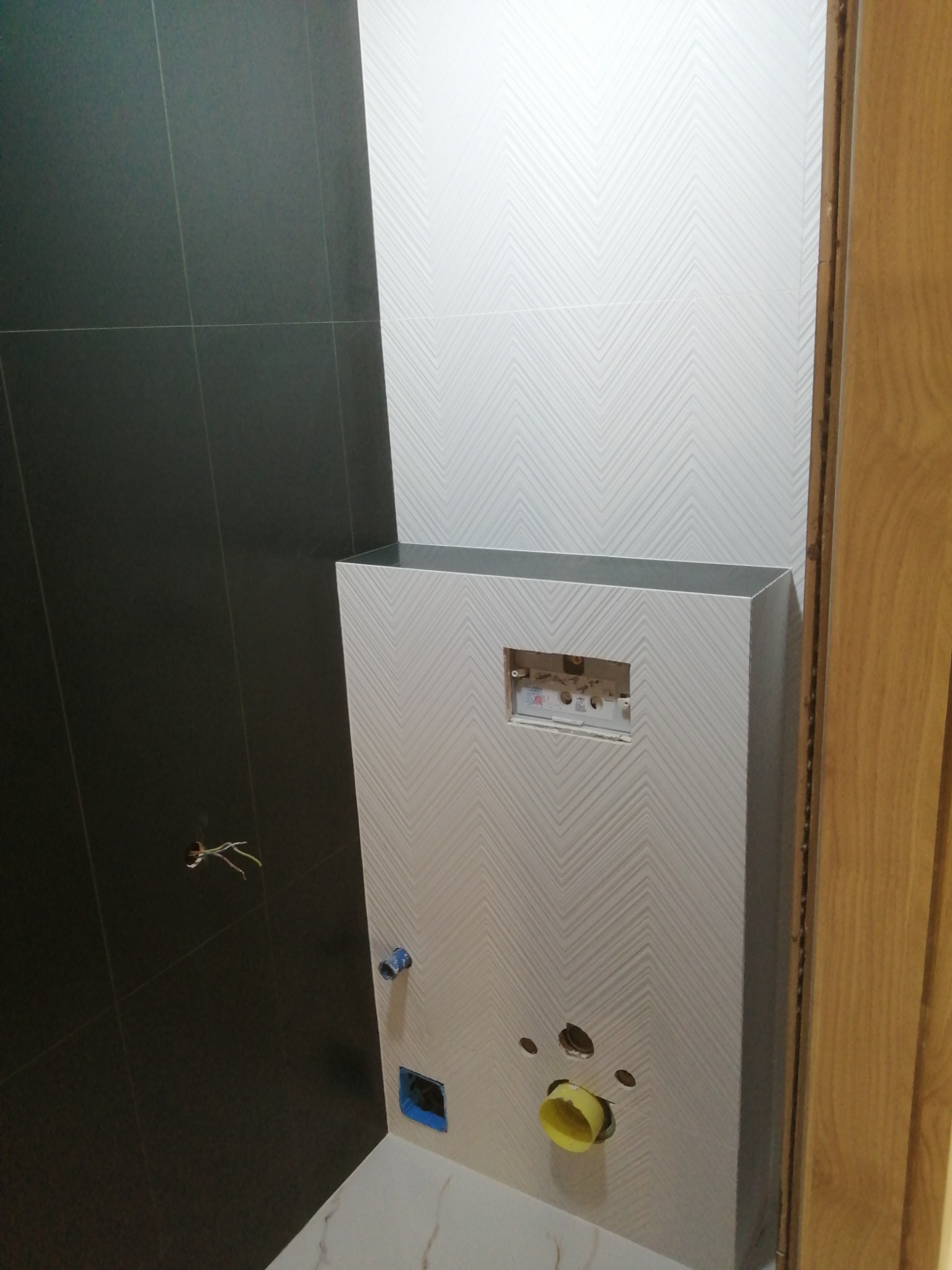 Wnętrze łazienki w trakcie prac: czarne płytki na ścianie, biała ściana z geometrycznym wzorem w jodełkę, przygotowane otwory na instalacje, widoczne kable elektryczne.