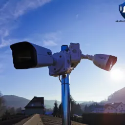 Dwie kamery monitoringu (BCS i Hikvision) zamontowane na metalowym maszcie na tle błękitnego nieba i zabudowań w oddali, z widocznym logo firmy alarmowo24.pl w prawym górnym rogu.