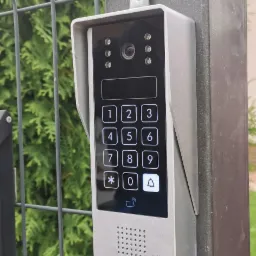 Szary wideodomofon Eura z klawiaturą numeryczną i kamerą zamontowany na metalowym słupku ogrodzenia panelowego, z zielenią w tle.