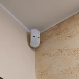 Biały czujnik ruchu alarmu zamontowany w rogu pomieszczenia, na styku gładkiej ściany i ściany z dekoracyjną strukturą.